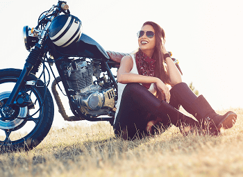 Woman sitting in a grassy field leaning against her motorcycle